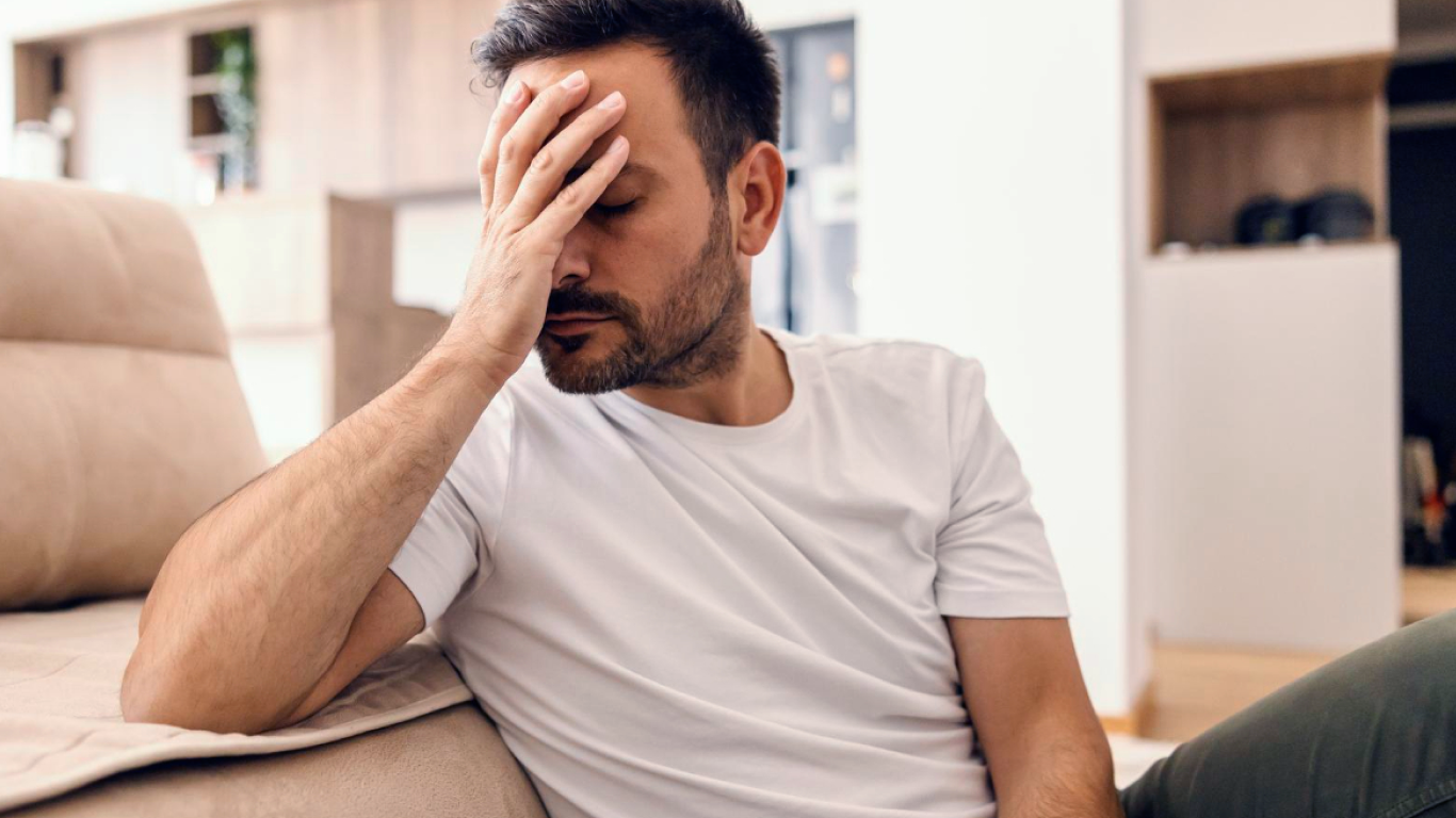 Homem no sofá, com a mão no rosto, sofrendo com o pênis torto