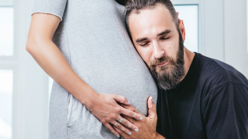 Homem com a cabeça reclinada na barriga de sua mulher gestante, feliz por recuperar a fertilidade masculina